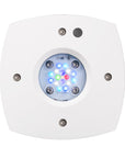 The AQUA Illumination Prime 16HD REEF is a white, square device with a circular LED display showing blue, green, and red lights. Its corners have metallic screws and a small circular component is located above the vibrant display.