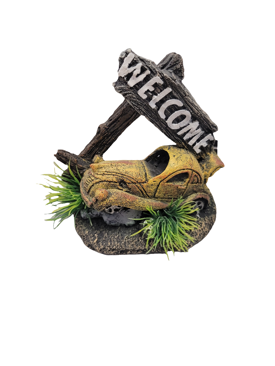 The Car with Welcome Sign Decor - 4.5in is a small sculpture showcasing a yellow, weathered car partially buried in dark soil with green plants. A wooden post featuring a diagonal WELCOME sign leans against the car on a white background.