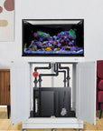 A stylish room features an Innovative Marine INT 112 Gallon Lagoon Aquarium Complete Reef System, showcasing a vibrant saltwater reef on a white cabinet. Open doors reveal plumbing and equipment. A wooden side table, abstract wall art, and red chair complete the decor.