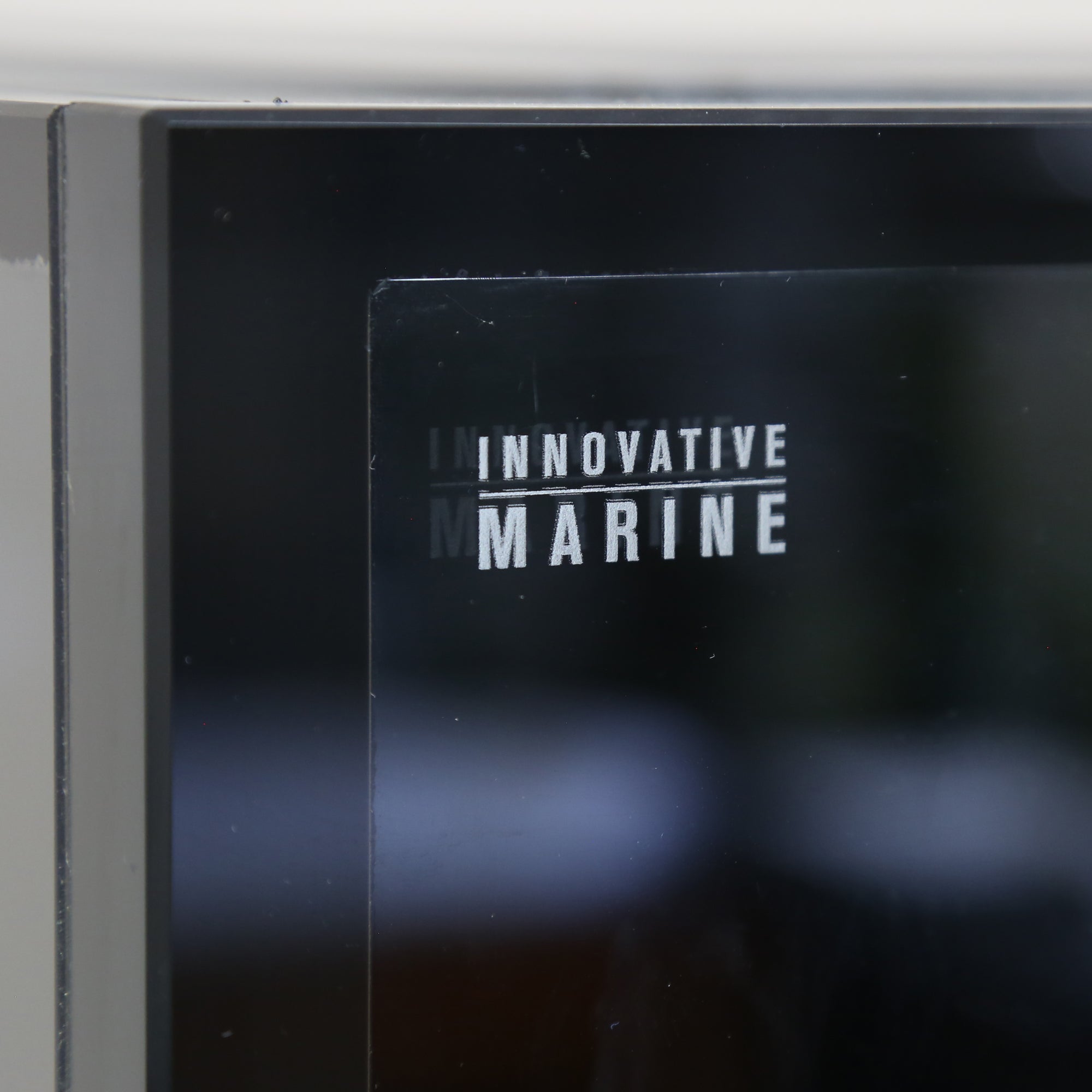 Close-up of a black glass panel featuring INNOVATIVE MARINE in white capitals. The reflection suggests its part of the Innovative Marine- EXT 200 Aquarium w/ APS Stand, designed for displaying your 200-gallon aquarium.