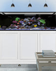 An Innovative Marine-EXT 200 Aquarium w/ APS Stand holds vibrant coral and fish atop its white cabinet. Three pendant lights illuminate the aquarium, while a small table is in front, and large windows are partially seen in the background.