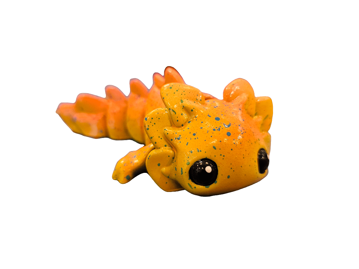 The AO Axolotl Trinket/Charm UV SML is a small, orange figurine with a cartoon style featuring black speckles and large black eyes, lying on a white background. Its whimsical design boasts exaggerated features.