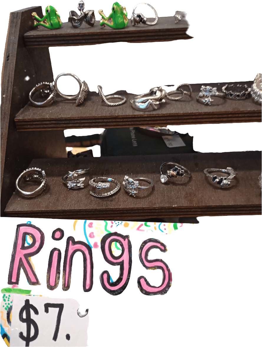 A wooden display stand showcases AO Rings, with a variety of designs and small stones. Two green frog figures rest on top of the stand, while a handwritten sign below announces: AO Rings $7.