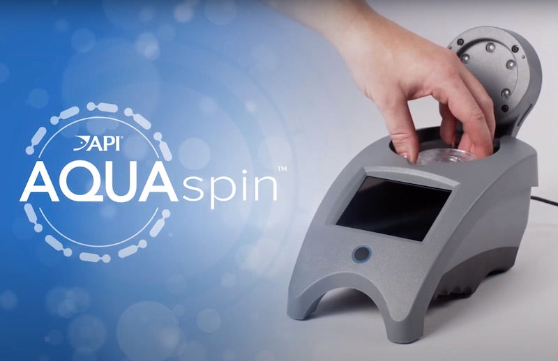 A hand is placing a water sample into a grey device with a screen labeled API AquaSpin - Water Test - SW & FW. The background showcases a circular logo design with the text API AquaSpin on a blue gradient backdrop.