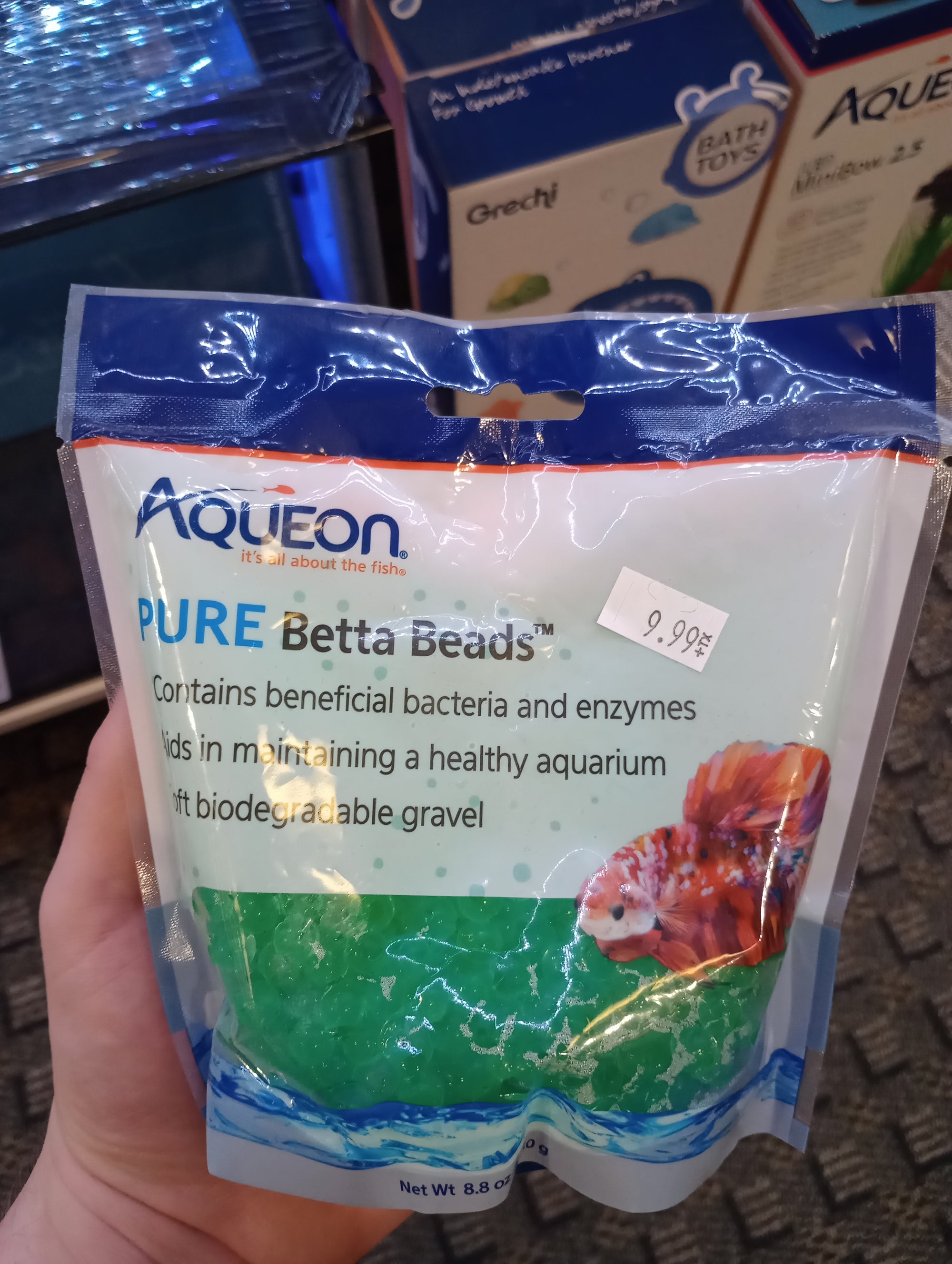 A hand holding a bag of Aqueon PURE Betta Beads Green, which contains beneficial bacteria and enzymes for promoting aquarium health. The front displays a price tag of $9.99.