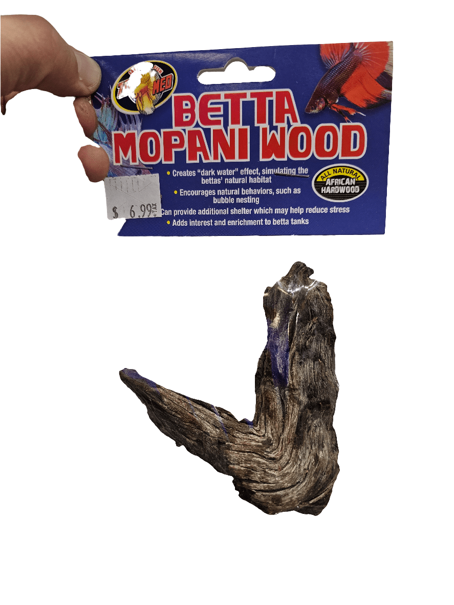 A hand holds a package labeled "ZooMed - Betta Mopani Wood l," priced at $6.99, ideal for betta tanks. Described as natural African hardwood, it creates dark water effects that encourage natural behaviors in betta fish. The wood piece is dark and irregularly shaped.